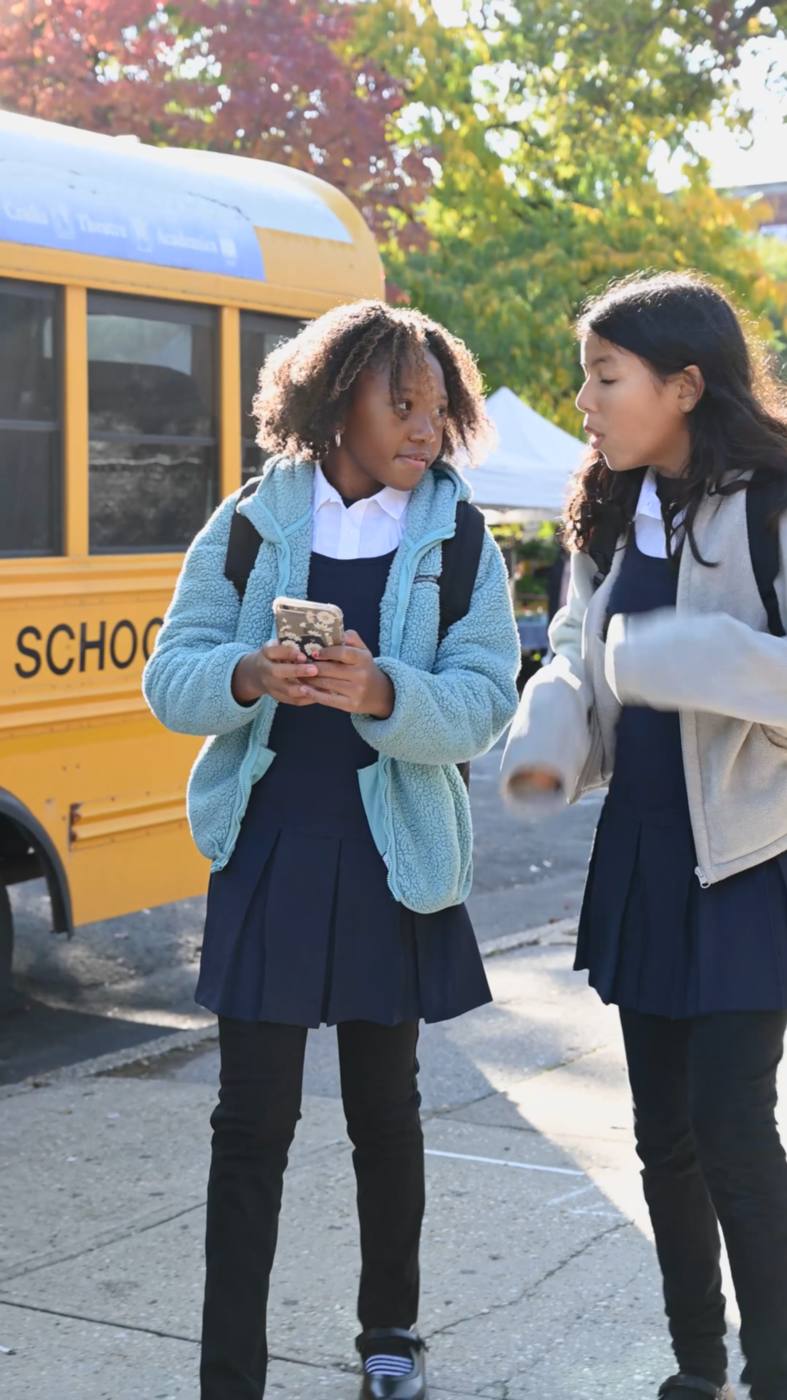 Two pupils in school uniform walking and talking together outside school, one holding a phone