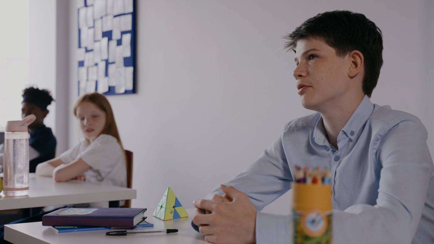 A secondary pupil sitting forward at a classroom table, listening, with two classmates working behind him