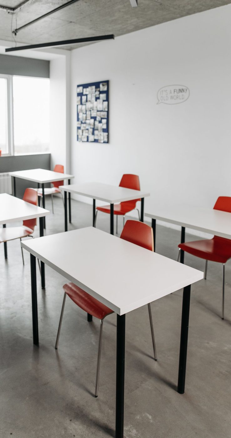 An empty classroom with white tables and red chairs, a display board of photographs on the wall