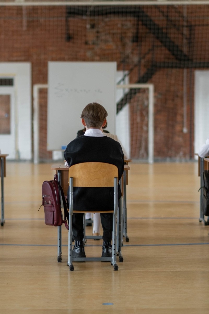 A boy alone at a single desk in a school hall laid out for an examination, a whiteboard behind him and empty floor all around
