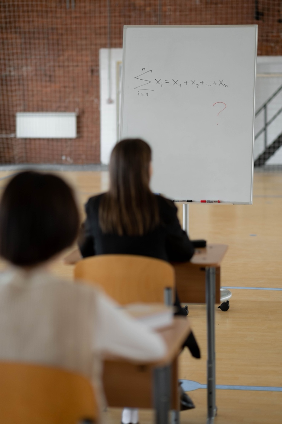 A whiteboard at the front of an examination hall carrying a summation formula and a red question mark, pupils seated in front of it