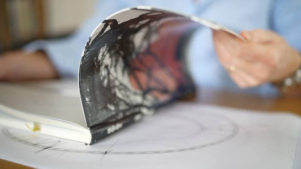 A hand turning the pages of a hardback book resting on a sheet of hand-drawn planning diagrams