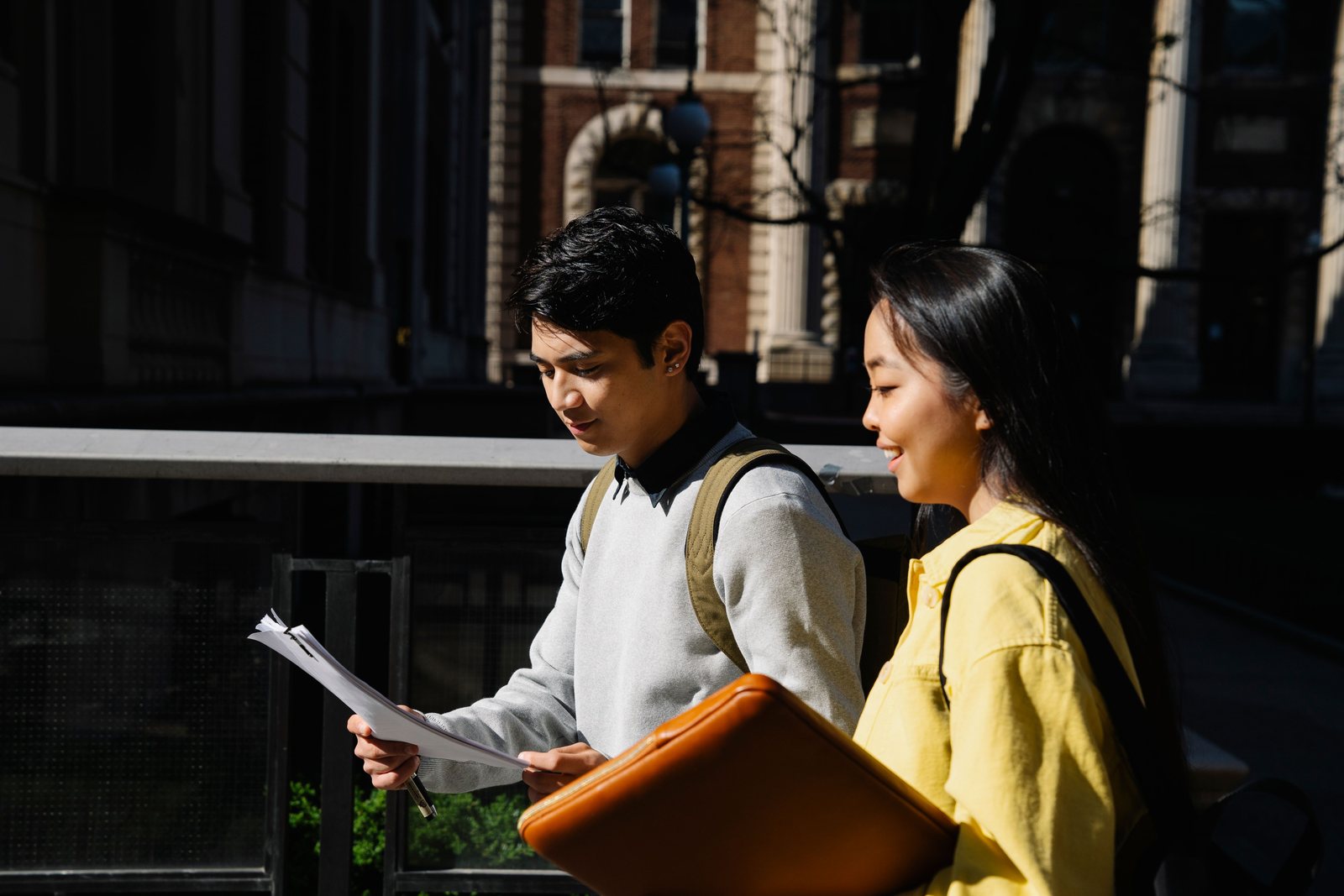 Two sixth-form students standing outside a school building in the sun, reading the papers in their hands