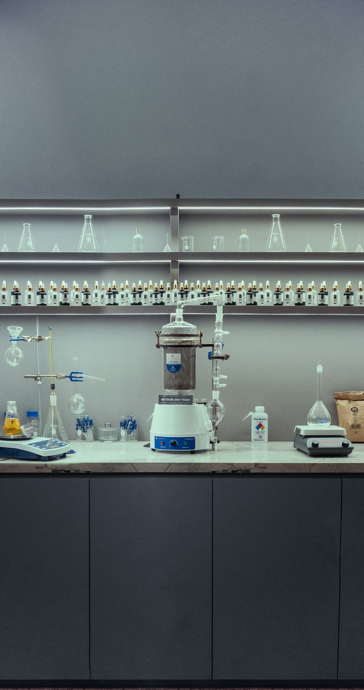 A laboratory bench with distillation apparatus, clamp stands and balances, under lit shelves lined with conical flasks and reagent bottles