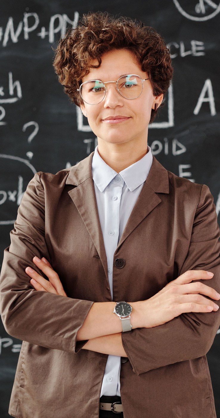 A teacher in a blazer standing with her arms folded in front of a blackboard, looking steadily at the camera